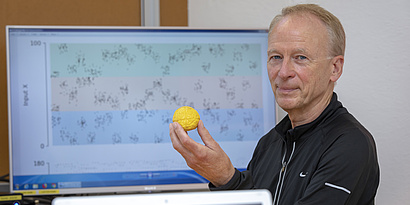 A man sits in front of a computer screen holding a small brain model in his hand, smiling at the camera.