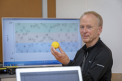 A man sits in front of a computer screen holding a small brain model in his hand, smiling at the camera.