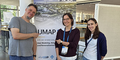 One male and two female researchers standing in front of a UMAP roll-up at the conference and smiling.