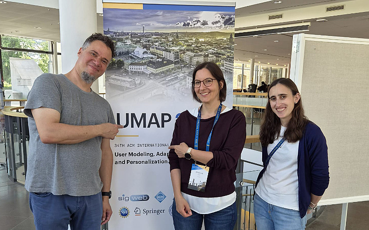 One male and two female researchers standing in front of a UMAP roll-up at the conference and smiling.
