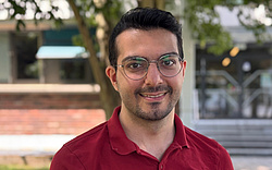 A man, Ali Kookani, smiling into the camera, wearing a red polo shirt and glasses.