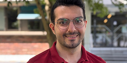 A man, Ali Kookani, smiling into the camera, wearing a red polo shirt and glasses.