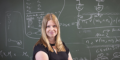 Bettina Könighofer smiles at the camera while standing in front of a blackboard covered with mathematical formulas.