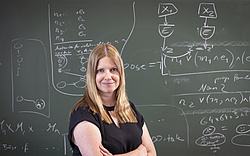Bettina Könighofer smiles at the camera while standing in front of a blackboard covered with mathematical formulas.